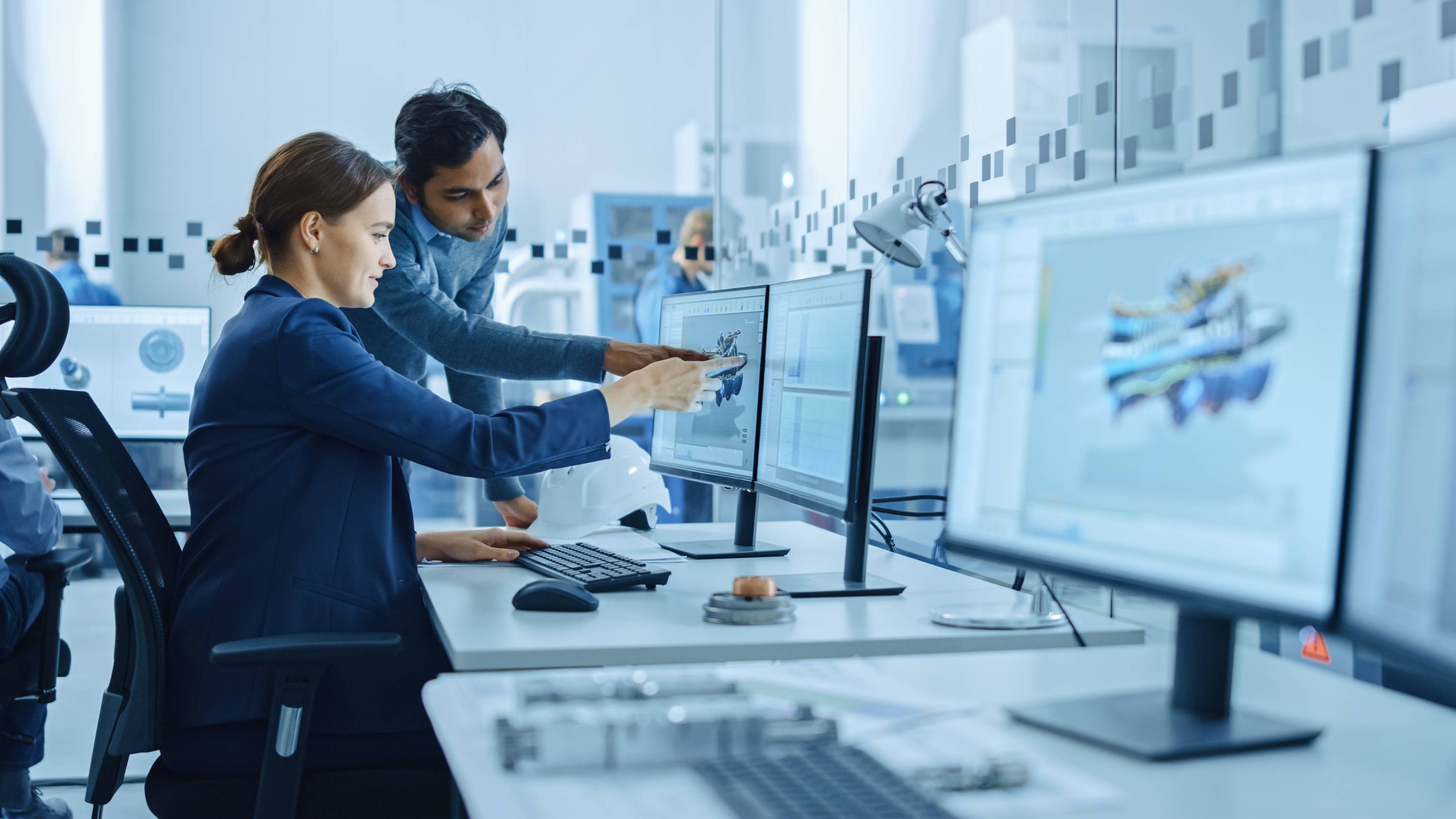 Two engineers collaborate at a computer, examining 3D models on dual monitors. The high-tech office setting conveys focus and innovation.
