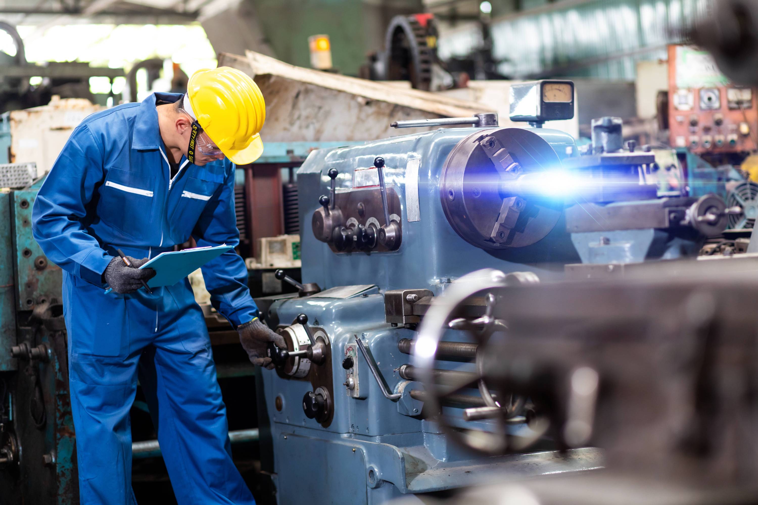 A worker in a blue jumpsuit and yellow helmet inspects a large industrial machine in a factory, holding a clipboard, focusing on machinery details.
